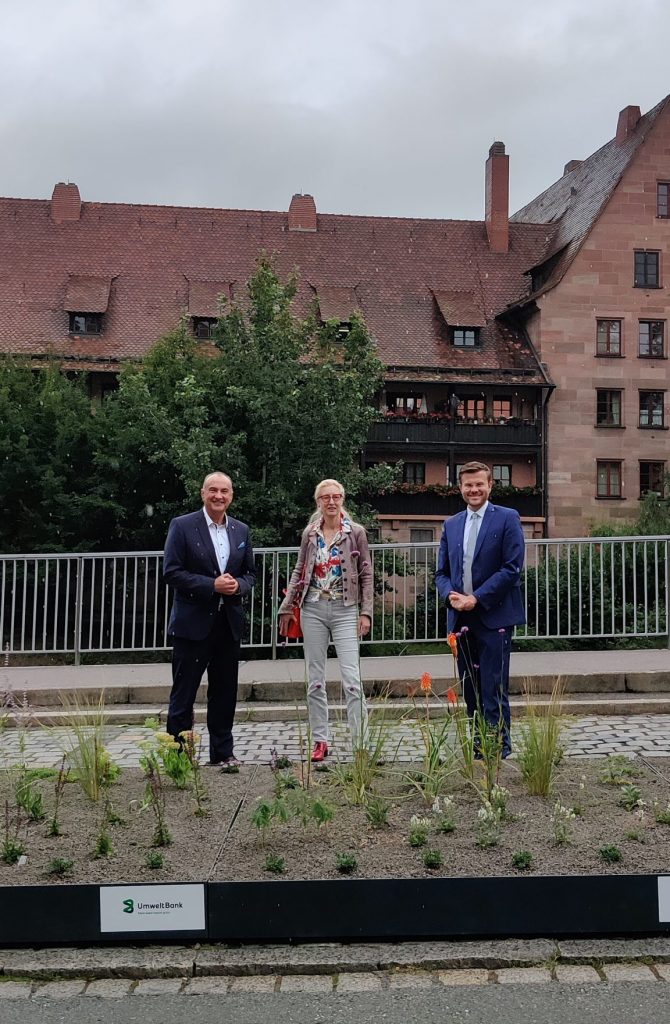 Goran Basic von der Umweltbank, Daniela Bock vom Verein Grünclusiv e.V. und Nürnbergs Oberbürgermeister Marcus König (von links nach rechts) stehen hinter dem mobilen Grün am Unteren Bergauerplatz.